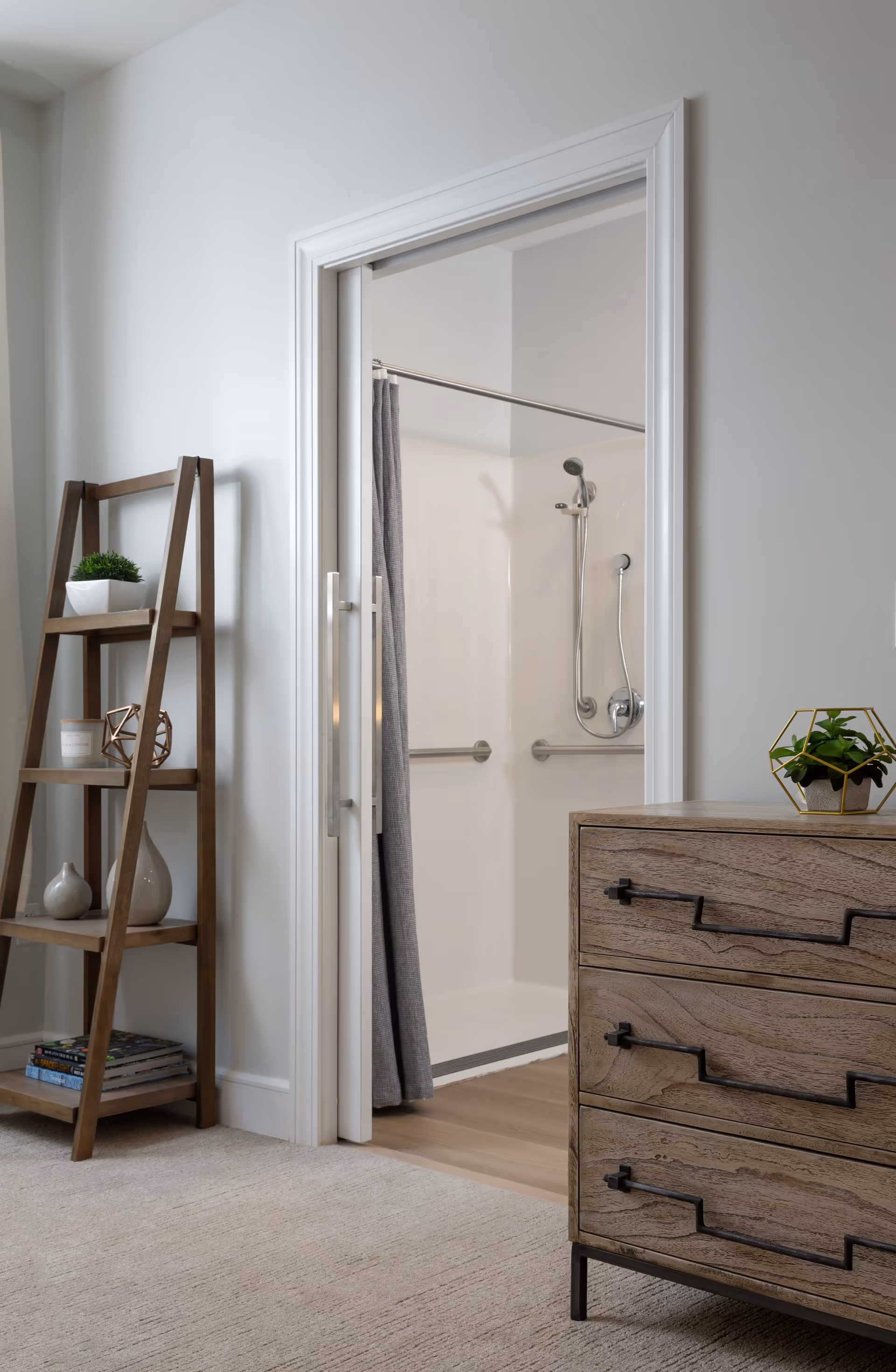 Open doorway reveals an accessible walk-in shower with grab bars and a gray curtain, next to a wooden dresser and ladder shelf.
