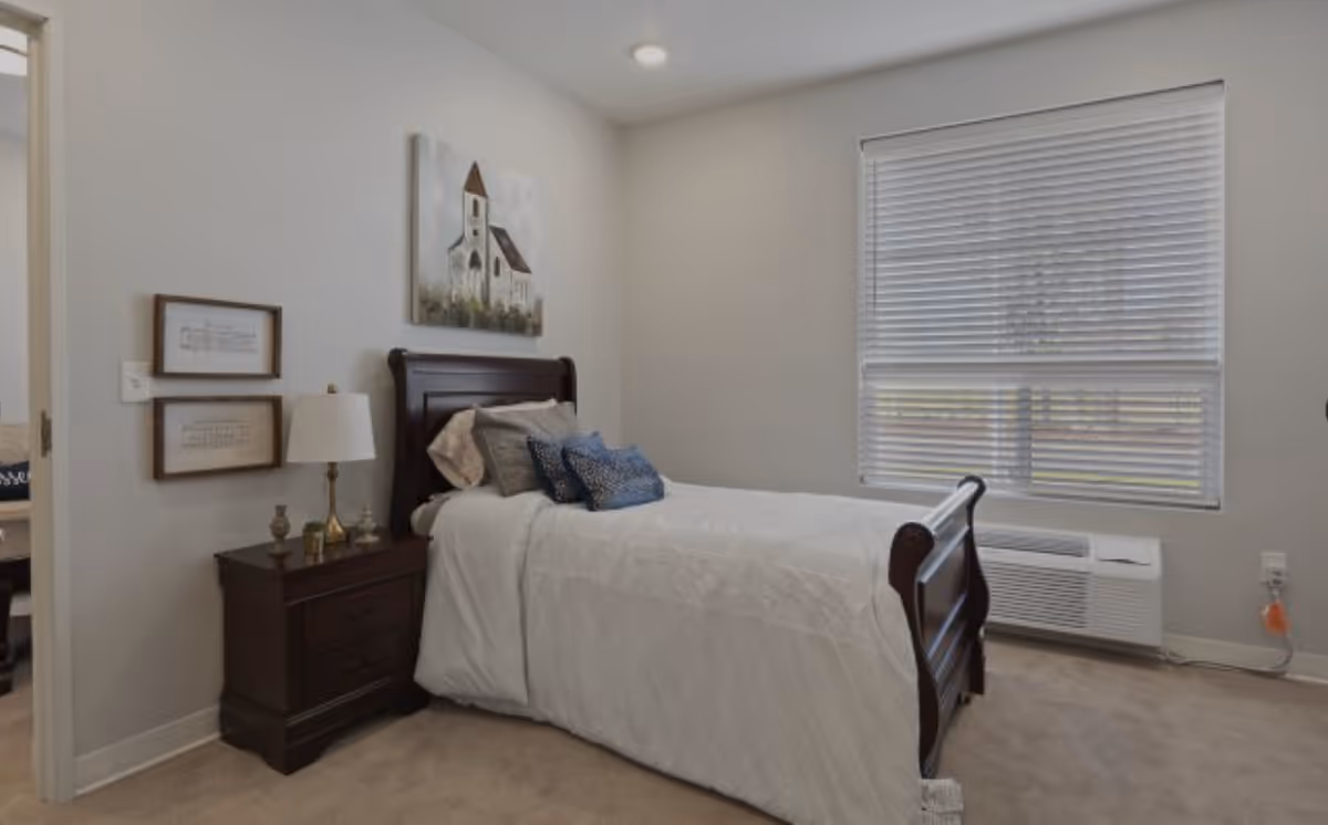A bedroom with a single dark wooden bed frame, white bedding, and several pillows. There is a dark wooden nightstand with a lamp and decorative items next to the bed. On the wall above the nightstand are two framed pictures, and above the bed is a painting of a church. A window with closed blinds is on the right side of the room, and a heating/cooling unit is below the window.