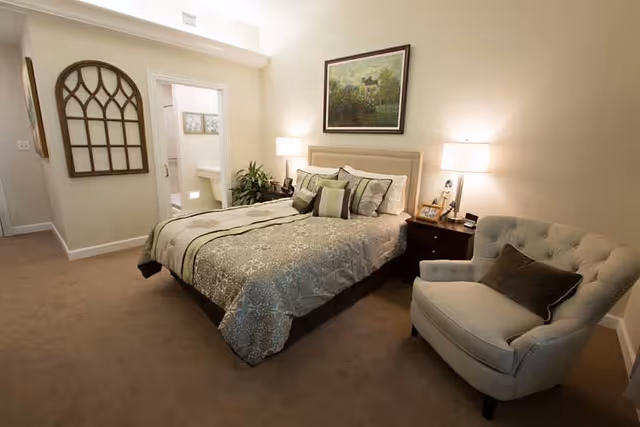 A cozy bedroom with a neatly made bed featuring patterned bedding and multiple pillows. There is a cushioned armchair with a dark pillow to the right of the bed. Two bedside tables with lamps flank the bed. A framed landscape painting hangs above the headboard. A decorative window frame is mounted on the wall to the left, and an open door reveals a bathroom with a sink and mirror.