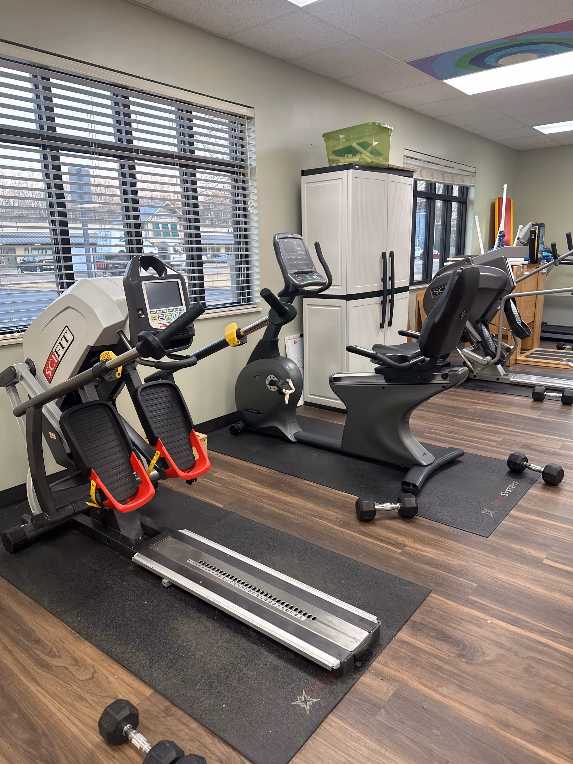 Indoor fitness room with exercise equipment including a rowing machine, stationary bike, and recumbent bike. The room has large windows with blinds, wooden flooring, and a white storage cabinet. Dumbbells are placed on the floor near the exercise machines.