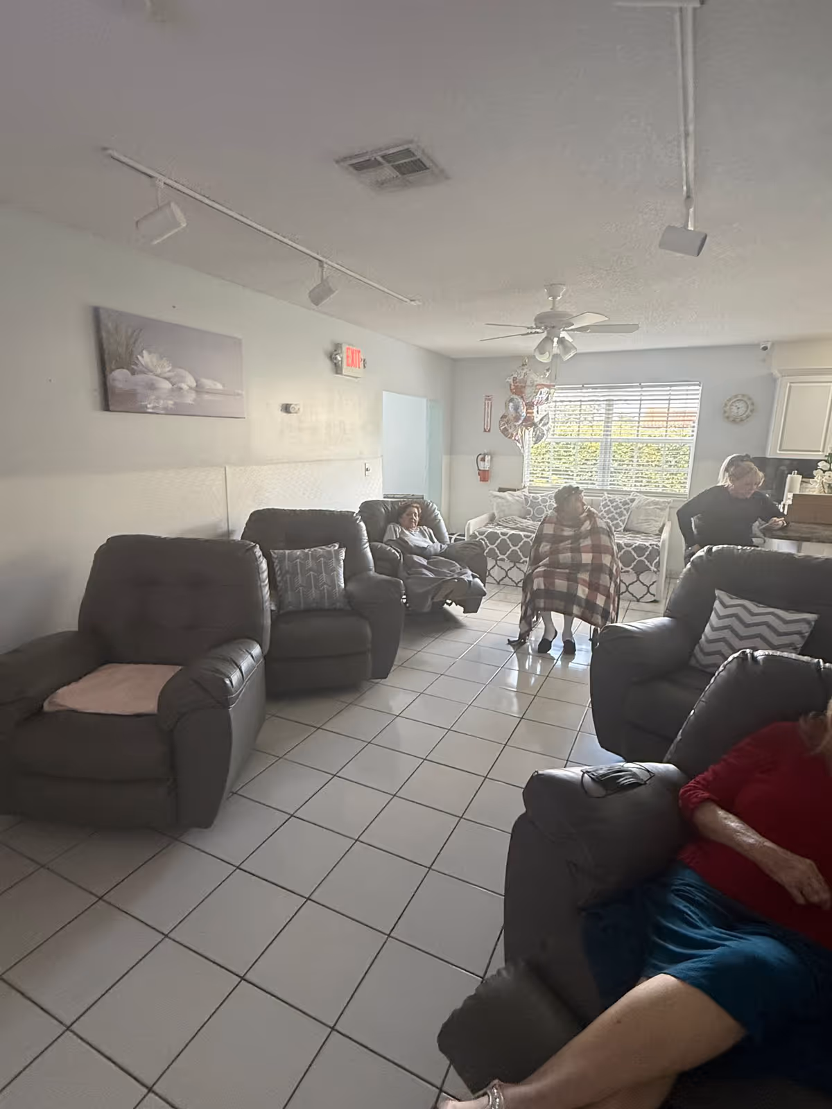 A living room area in a senior living facility with several comfortable recliner chairs arranged on a tiled floor. There are four elderly individuals present, one wrapped in a blanket, and another sitting on a patterned couch near a window with blinds. The room has a ceiling fan, a wall painting, and an exit sign above a door.