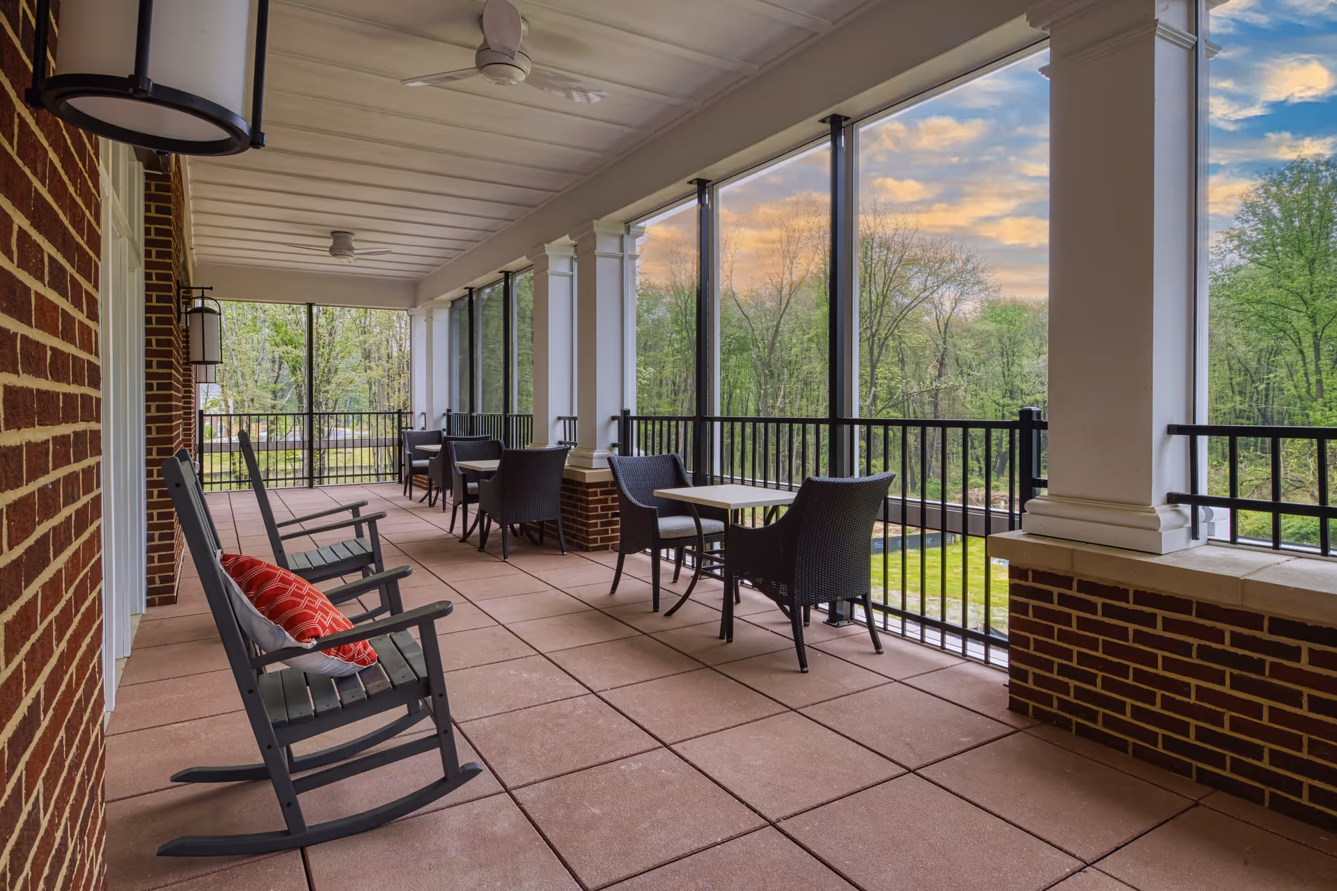 A covered outdoor patio area with rocking chairs and tables with chairs, overlooking a green landscape with trees under a partly cloudy sky.