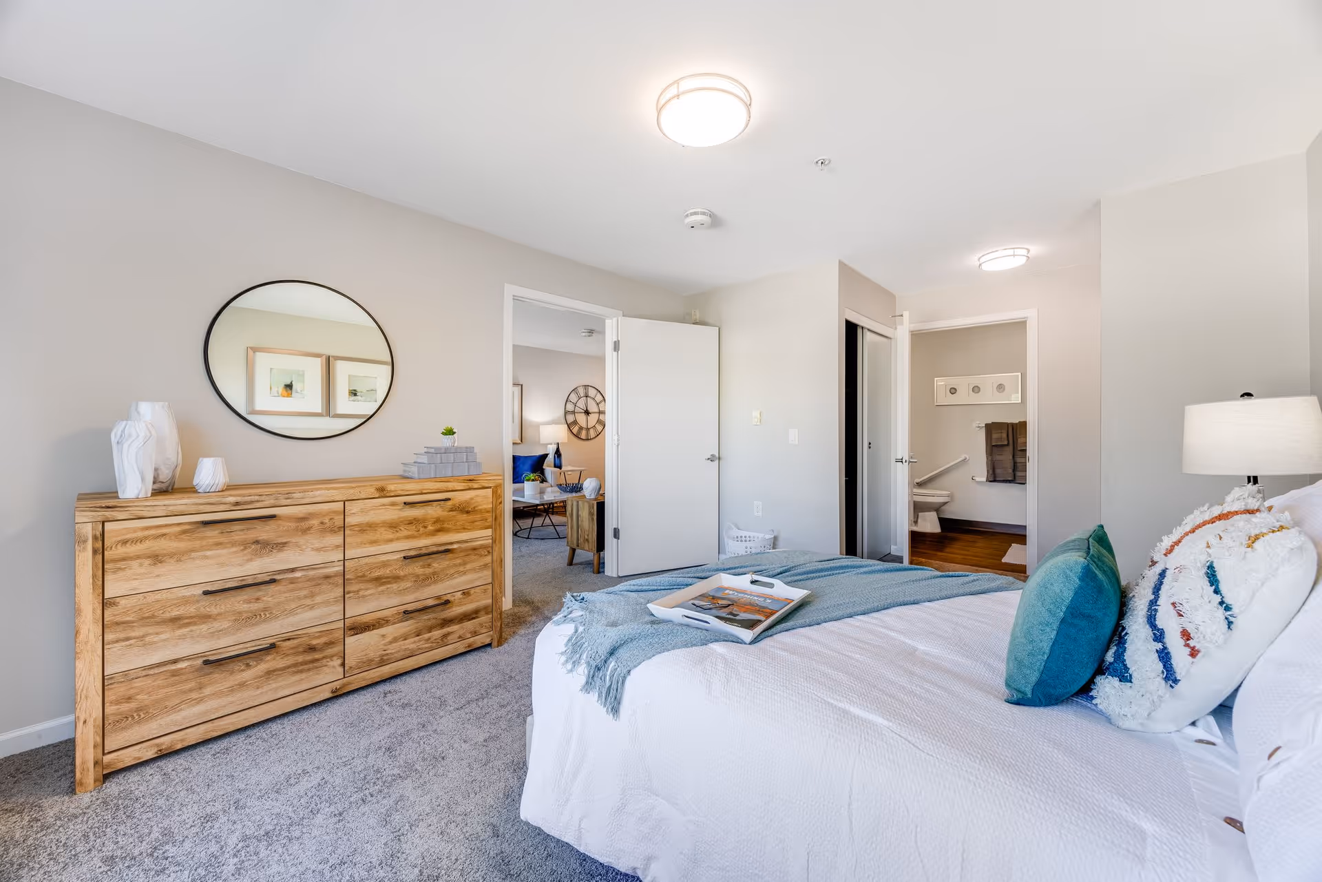 A bright bedroom with a neatly made bed featuring white bedding and colorful pillows. A wooden dresser with decorative items and a round mirror hangs on the wall above it. The room has light gray walls and carpeted flooring. An open door reveals a glimpse of a living area with a clock on the wall, and another door leads to a bathroom with a toilet and towels visible.