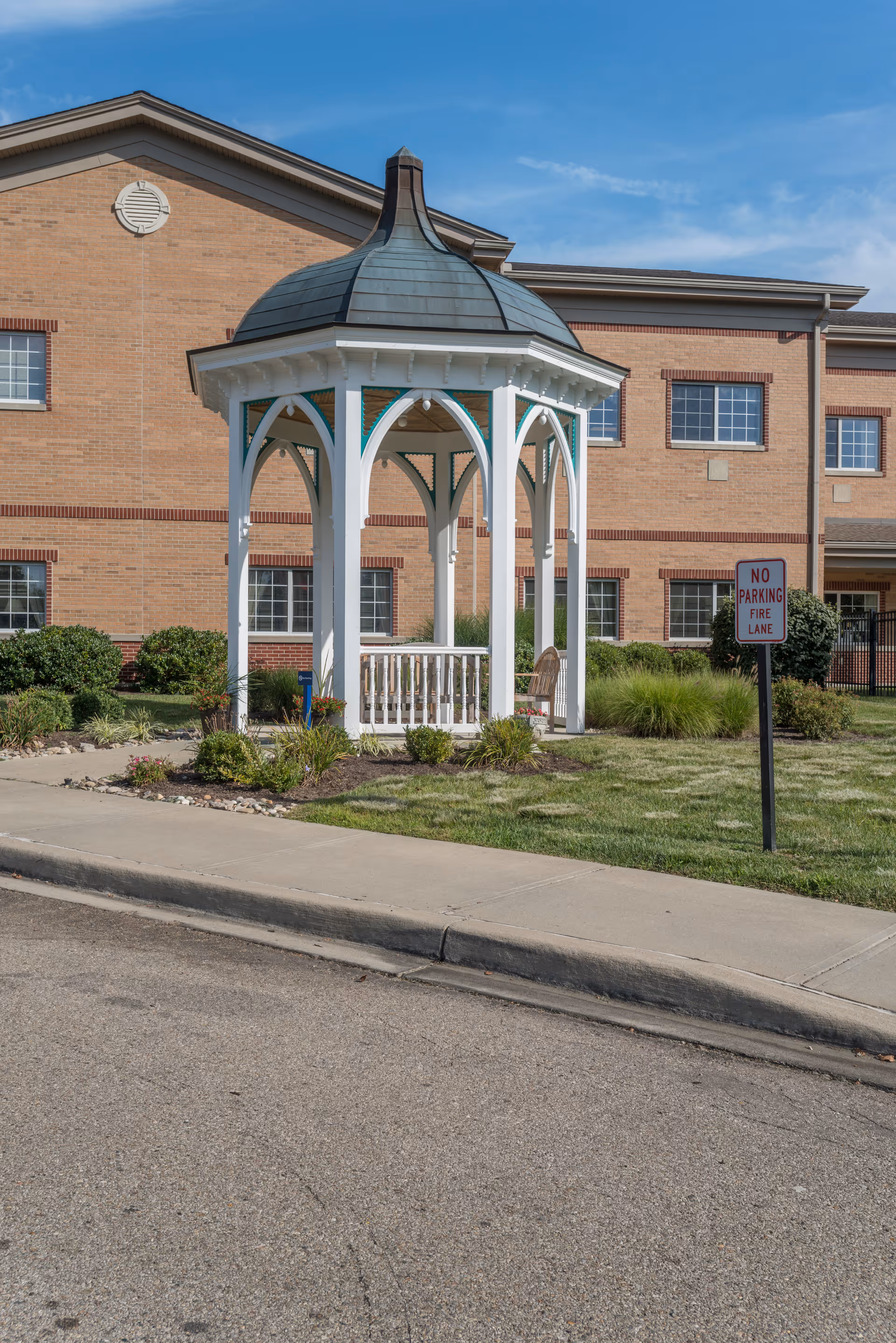 A white gazebo with a greenish-gray roof situated on a grassy area in front of a brick building under a clear blue sky. There is a 'No Parking Fire Lane' sign near the gazebo and some landscaping with bushes and plants around it.