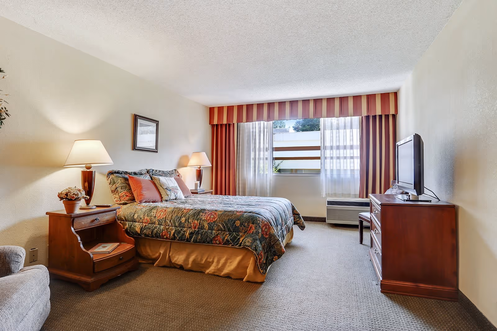 Neatly furnished bedroom with a double bed topped with a patterned bedspread, bedside tables and lamps, a TV on a dresser, and a window with striped valance.