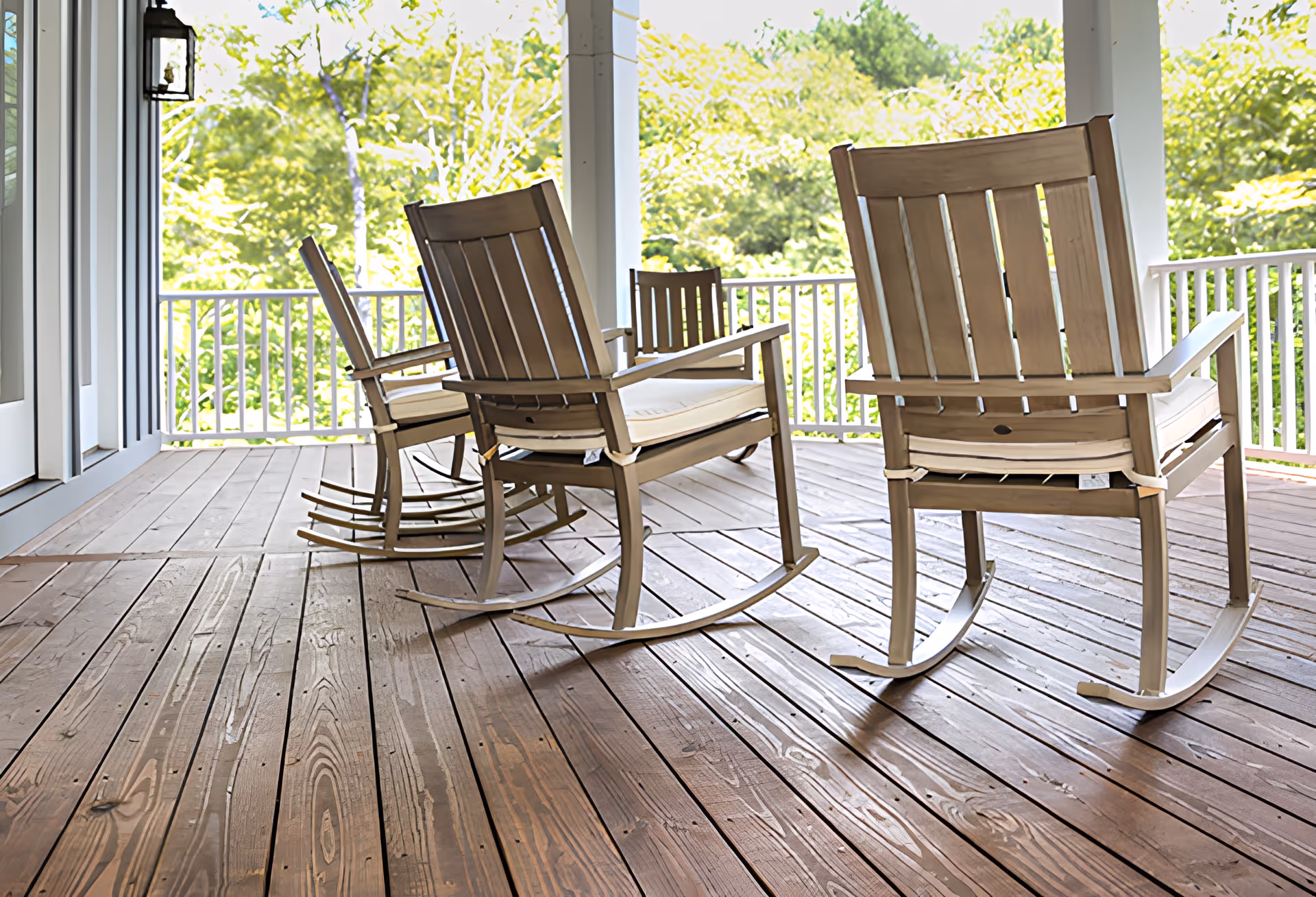 A wooden porch with three wooden rocking chairs with cushions, overlooking a green leafy outdoor area with trees. The porch has wooden flooring and white railings.
