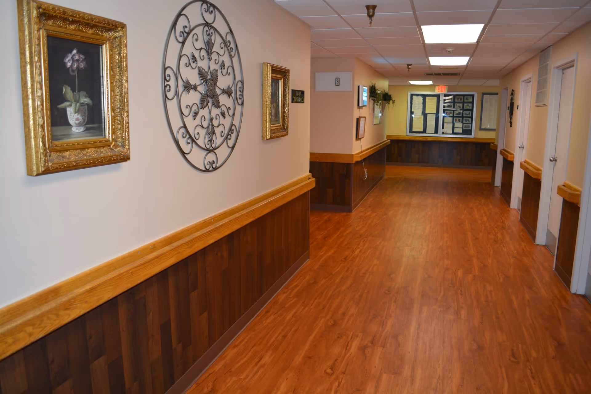 A clean, well-lit hallway with wooden flooring and wood paneling on the lower half of the walls. The walls are decorated with framed artwork and a decorative metal wall piece. Doors line the right side of the hallway, and a bulletin board is visible at the far end.