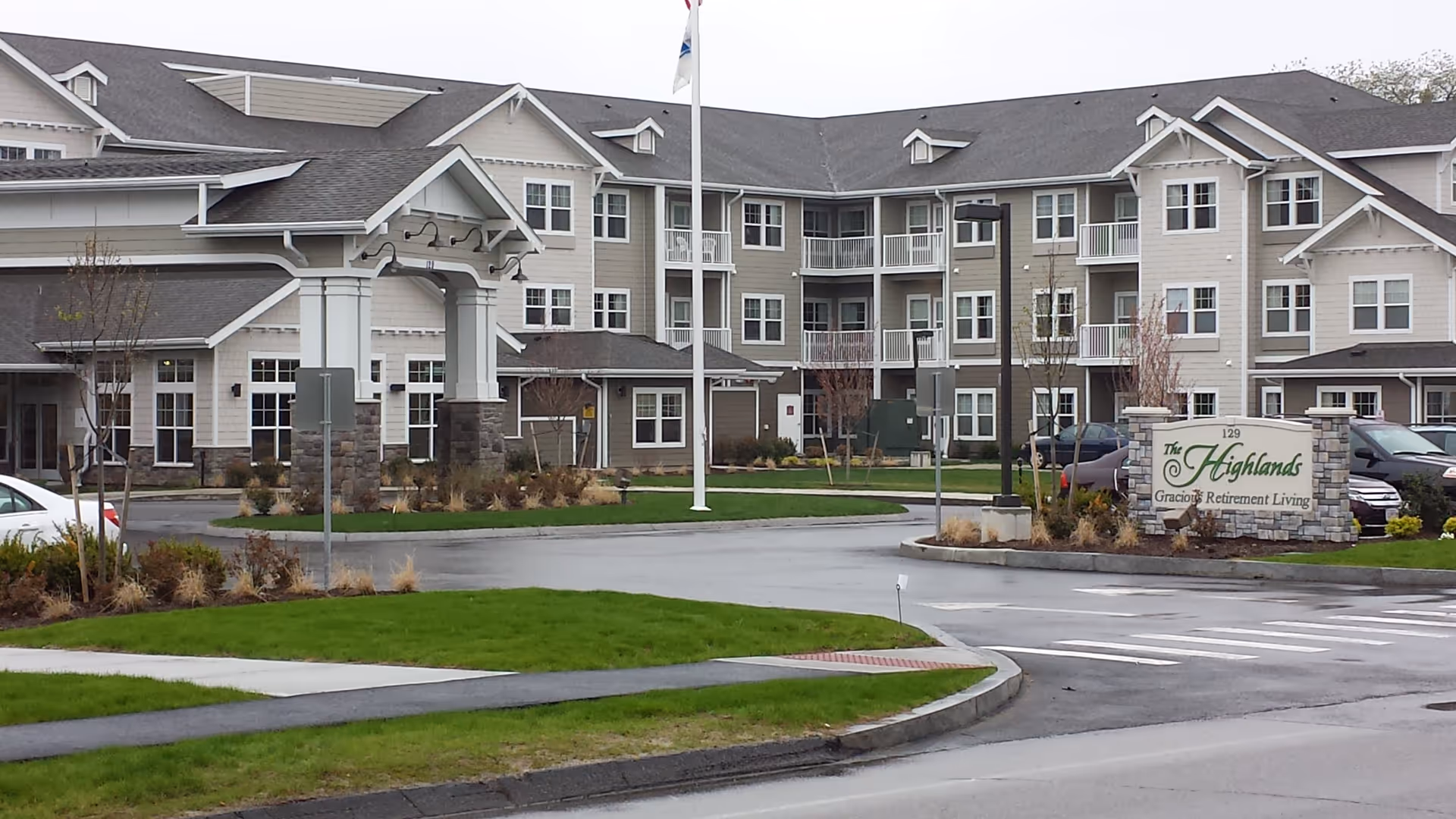 Exterior view of The Highlands Gracious Retirement Living facility showing a large multi-story building with multiple windows, a covered entrance, a flagpole, landscaped green lawns, and a sign with the facility name near the parking area.
