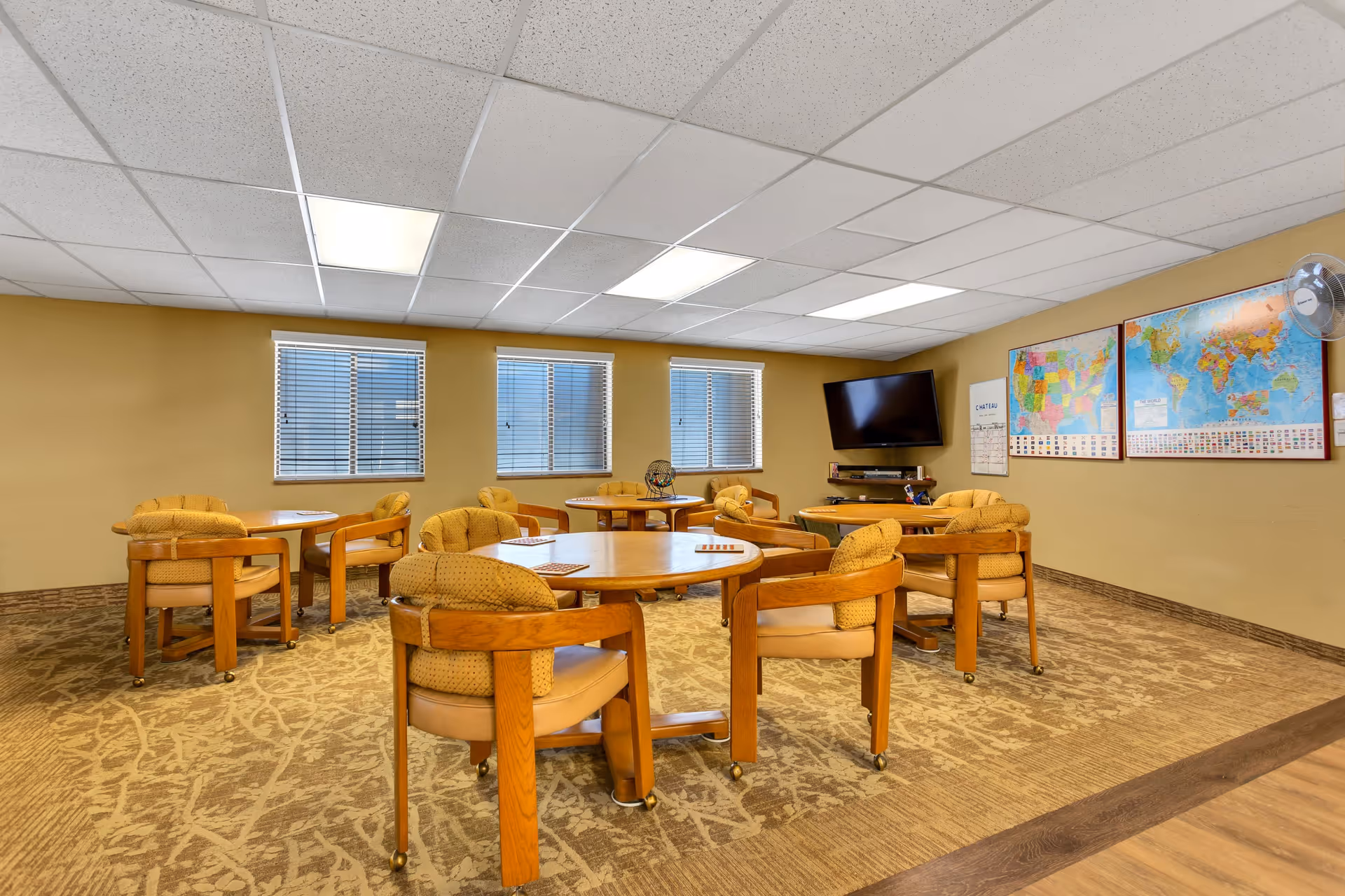 A senior living community activity room with round wooden tables and cushioned chairs on wheels. The room has beige walls, three windows with blinds, a wall-mounted flat screen TV, and two large maps on the wall. The carpet has a patterned design and the ceiling has white tiles with fluorescent lighting.