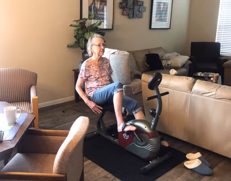 An older woman pedals a stationary exercise bike in a furnished living room area.
