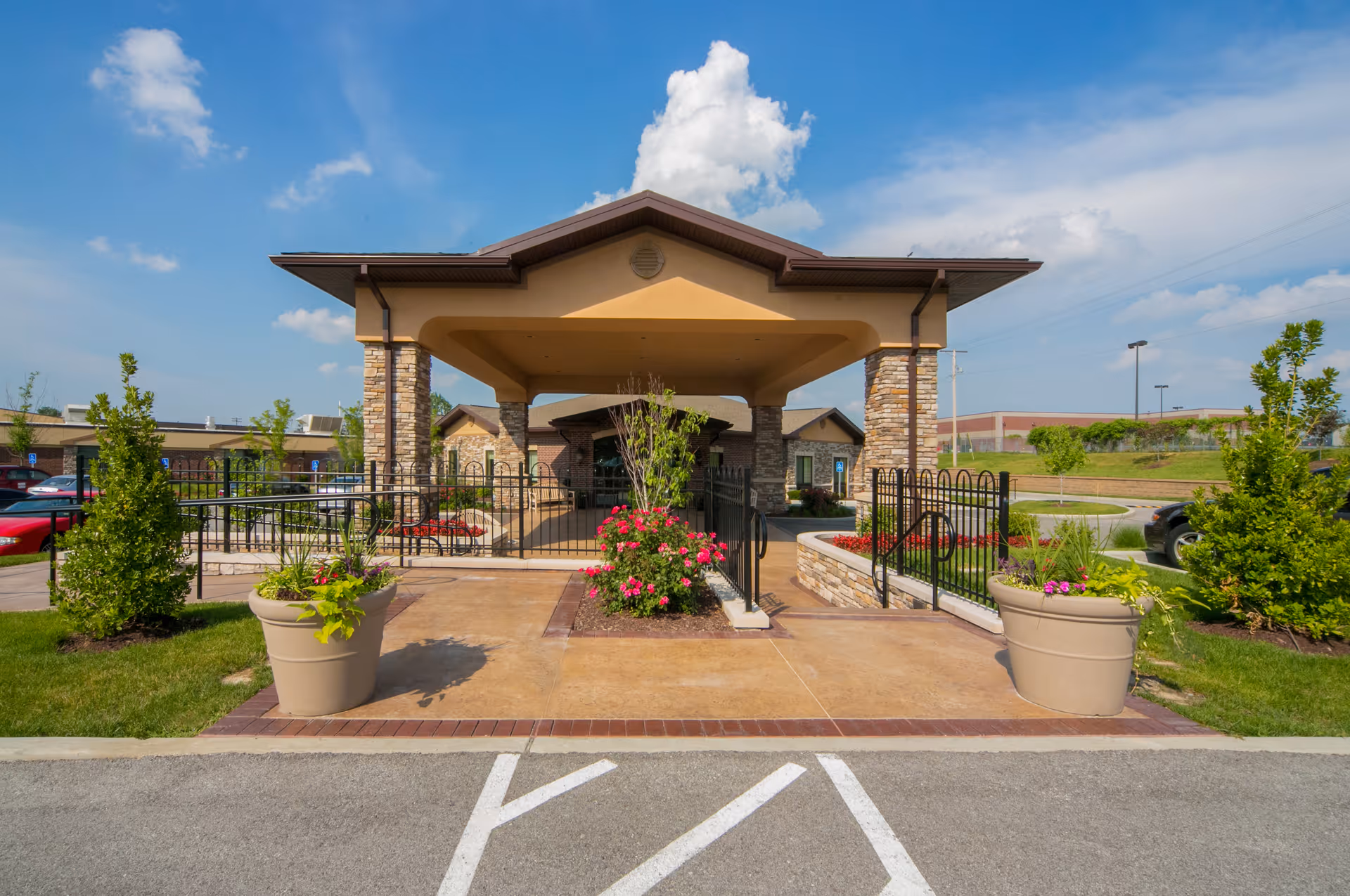 Front entrance of Life Care Center of Bridgeton featuring a covered drop-off area with stone pillars, landscaped flower beds, large planters with greenery, and a clear blue sky with some clouds.