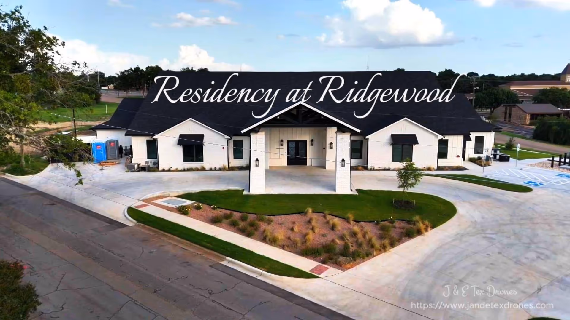 Front exterior view of a single-story building with a black roof and white walls, labeled Residency at Ridgewood. The building has a covered entrance supported by white pillars, surrounded by a landscaped area with grass and plants. There are parking spaces, including handicapped spots, visible on the right side.
