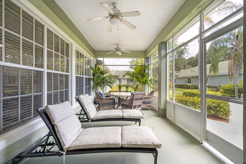 A screened-in porch area with two cushioned lounge chairs in the foreground and a round table with four wicker chairs in the background. The porch has ceiling fans and large windows overlooking a garden with palm trees and shrubs.
