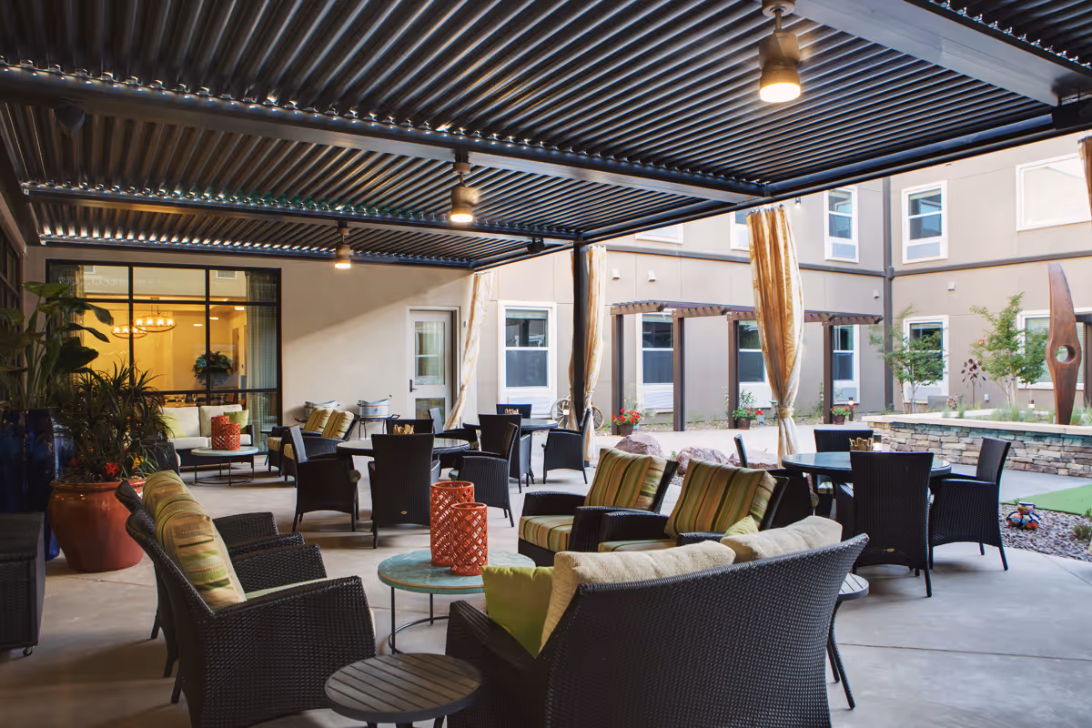 Covered outdoor patio area with wicker chairs and tables, some with cushions and pillows, under a metal pergola. The patio overlooks a courtyard with plants, a stone wall, and modern sculptures. The building windows and doors are visible in the background.