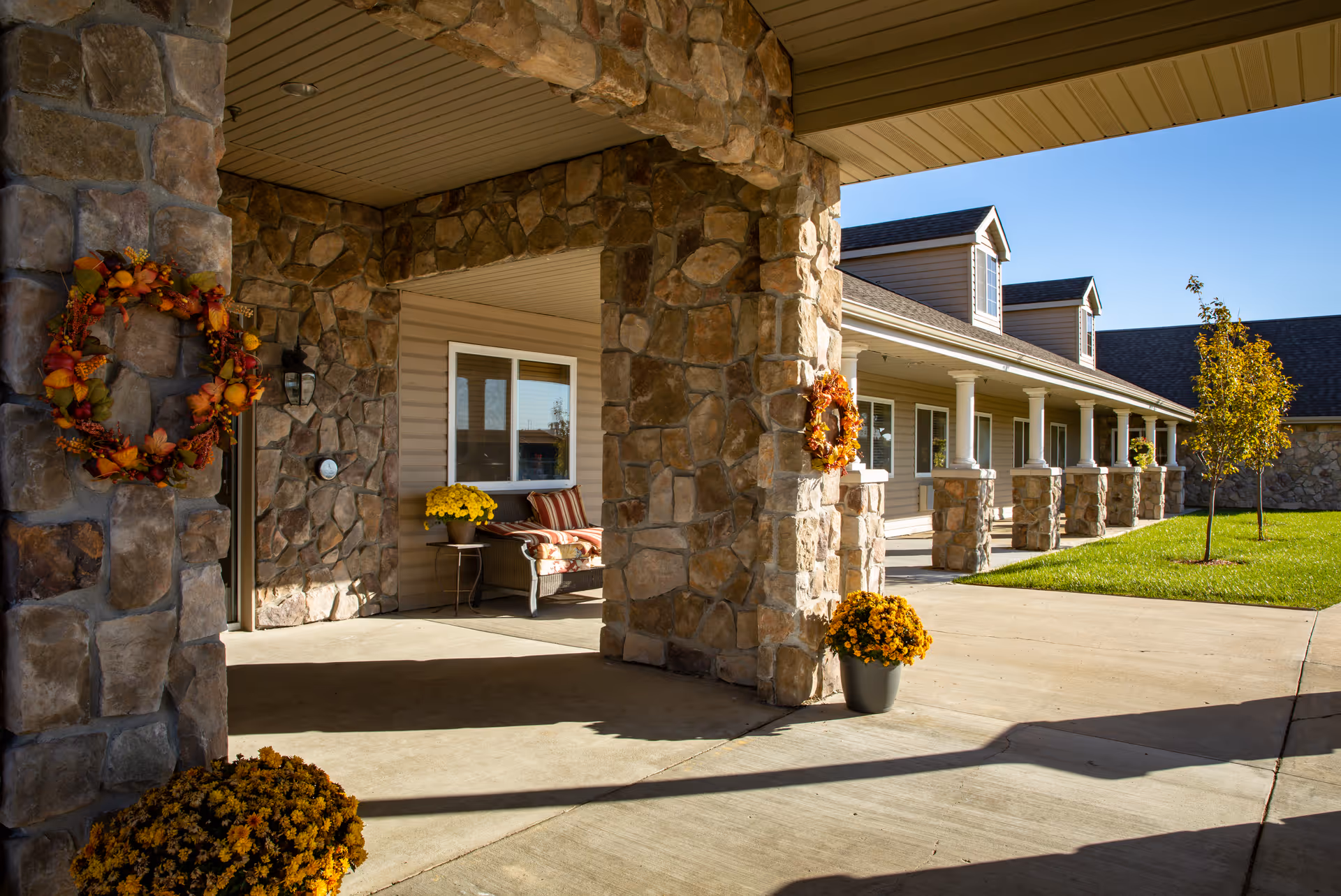Stone-column covered entrance and porch of an assisted living building decorated with autumn wreaths, potted mums, and a bench.