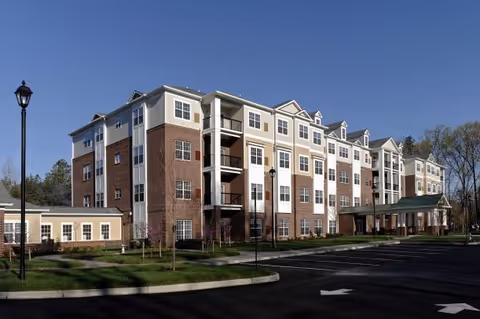 Front exterior of a multi-story senior living building with brick and light siding, balconies, a covered entrance, and an adjacent parking lot under a clear blue sky.