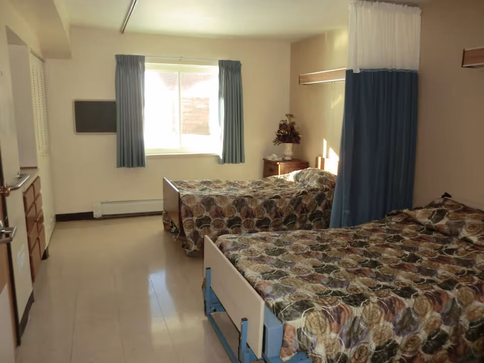 Shared resident bedroom with two hospital-style beds covered in floral bedspreads, a window with curtains, a nightstand, and a privacy curtain between beds.