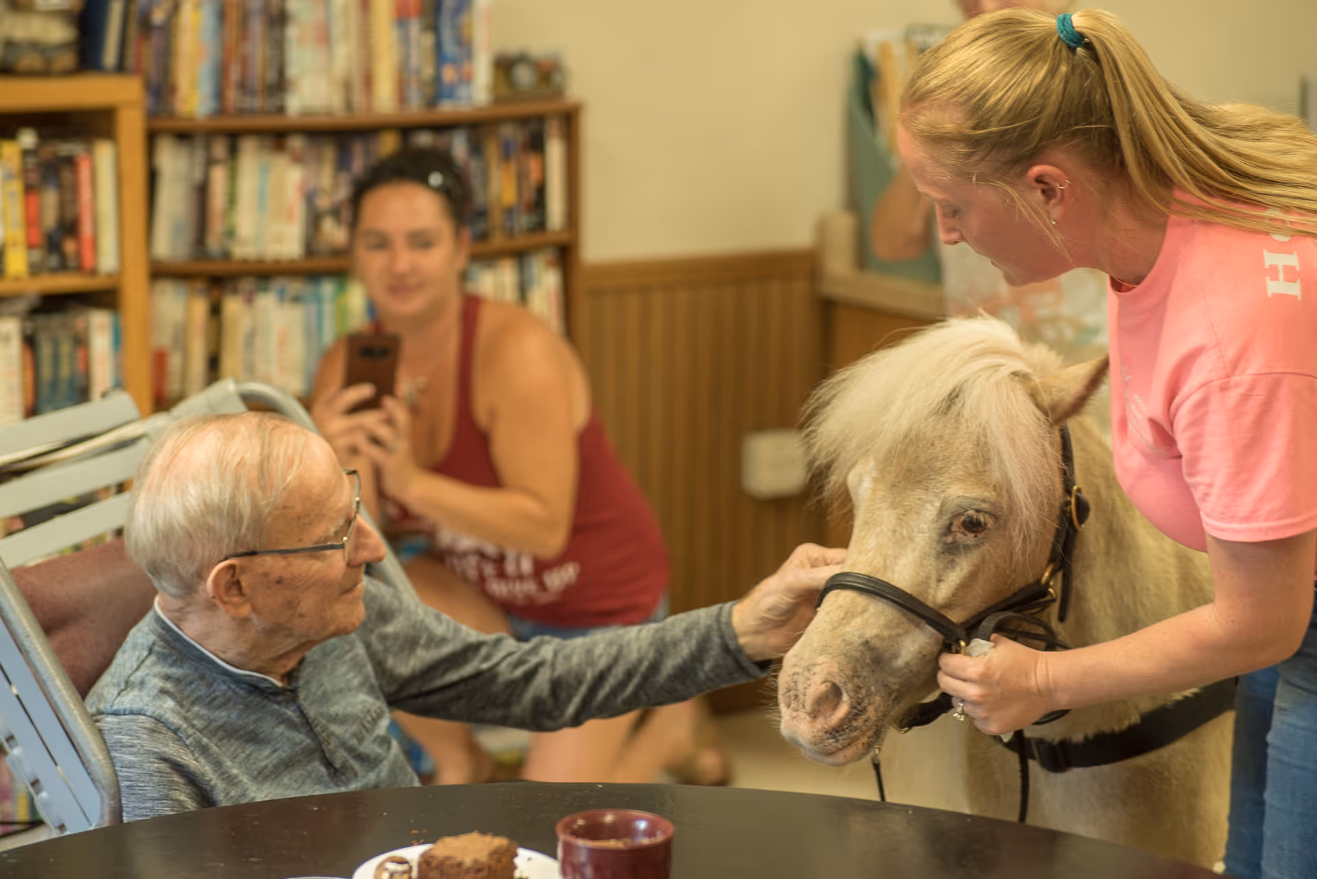 An elderly man sitting at a table inside a room with bookshelves in the background, reaching out to pet a small white pony held by a woman in a pink shirt, while another woman in a red tank top takes a photo with her phone.
