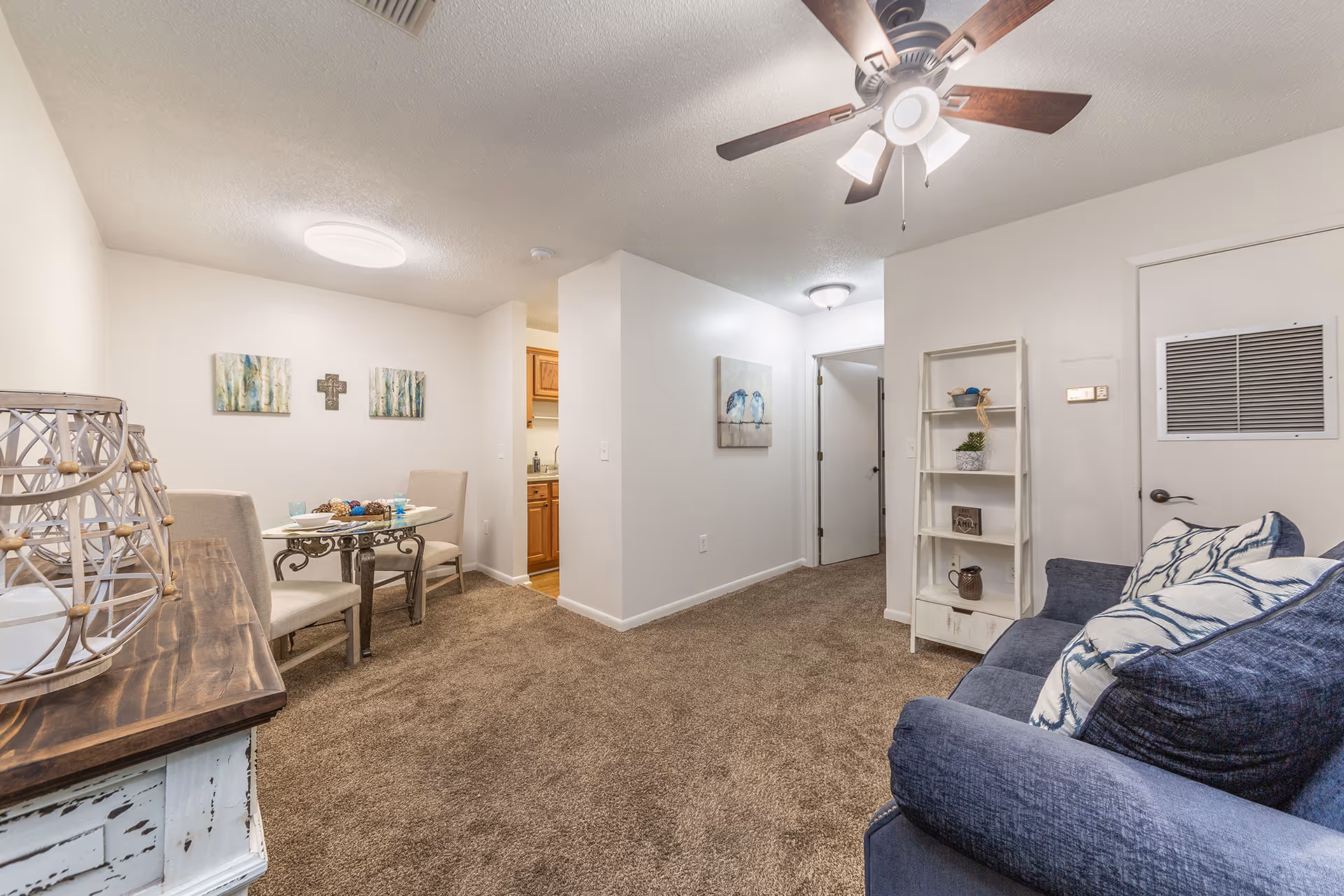 Interior view of a living area in Rosewood Assisted Living featuring a blue couch with patterned pillows, a white shelving unit with decorative items, a small glass dining table with two beige chairs, and a ceiling fan with lights. The room has beige carpet, white walls adorned with artwork, and a glimpse into a kitchen area with wooden cabinets.