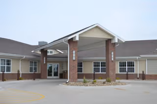 Entrance of a single-story retirement community building with a covered drop-off area supported by brick columns, beige siding, multiple windows, and a paved driveway.