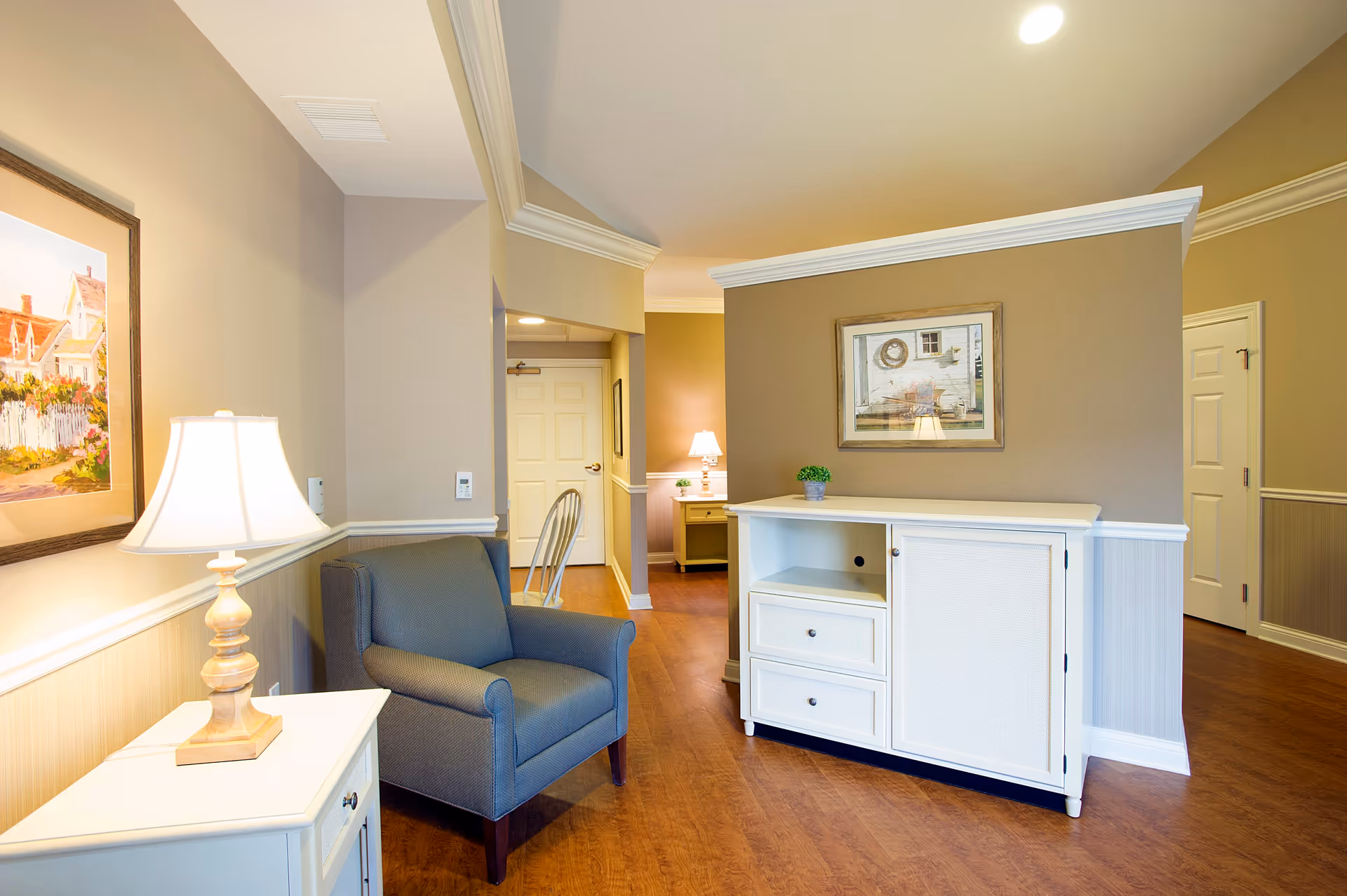 A cozy interior space with a gray armchair next to a white side table with a lamp. There is a white cabinet with drawers and a small green plant on top. The walls are painted beige with white crown molding and wainscoting. Two framed paintings hang on the walls, and the floor is wood.