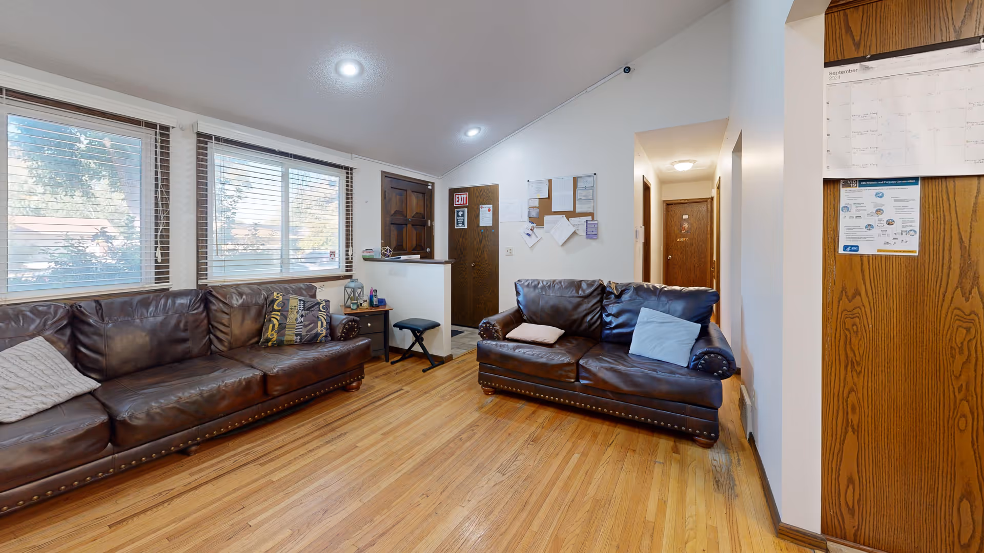 A cozy living room with two dark brown leather sofas, each with decorative pillows. The room has wooden floors and three large windows with blinds letting in natural light. A wooden front door is visible with an exit sign above it, and a bulletin board with papers is mounted on the wall near the door. There is a small side table with a lantern and some items on it, and a hallway leading to other rooms.