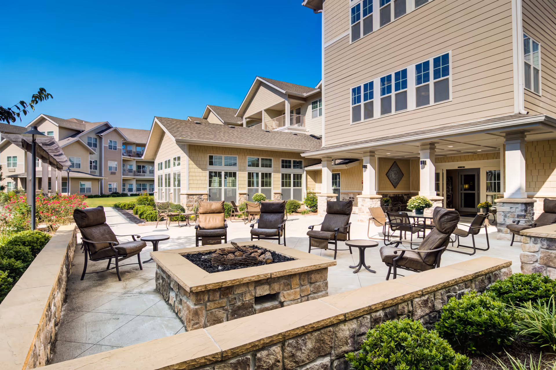 Outdoor patio area at Sanford Estates Gracious Retirement Living featuring cushioned chairs arranged around a stone fire pit, with additional tables and chairs under a covered porch. The building exterior is beige with multiple windows, and there are landscaped bushes and flowers surrounding the patio under a clear blue sky.