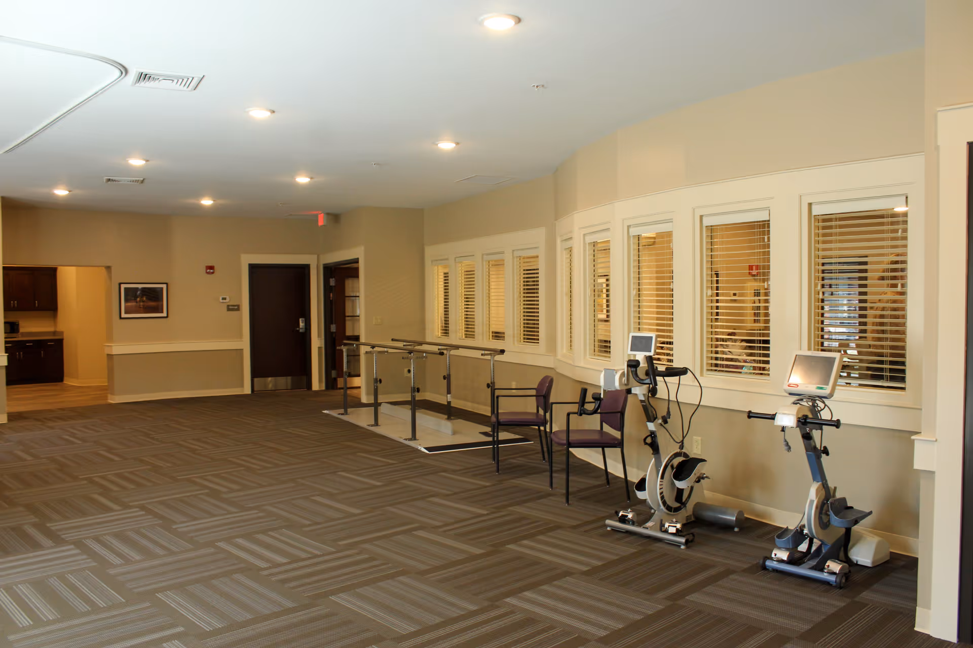 Interior view of a senior living facility hallway with exercise equipment including two stationary bikes and parallel bars for physical therapy. There are two chairs next to the equipment and windows with blinds on the right wall. The area is well-lit with ceiling lights and has patterned carpet flooring.