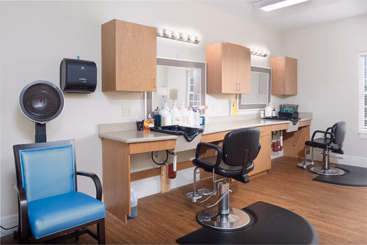 Interior view of a salon area with two black salon chairs in front of a counter with sinks and mirrors. There are wooden cabinets mounted on the wall above the counter, various hair care products on the counter, a blue chair, and a hair dryer hood on the left side. The floor is wooden, and the room is well-lit with natural light coming through a window on the right.