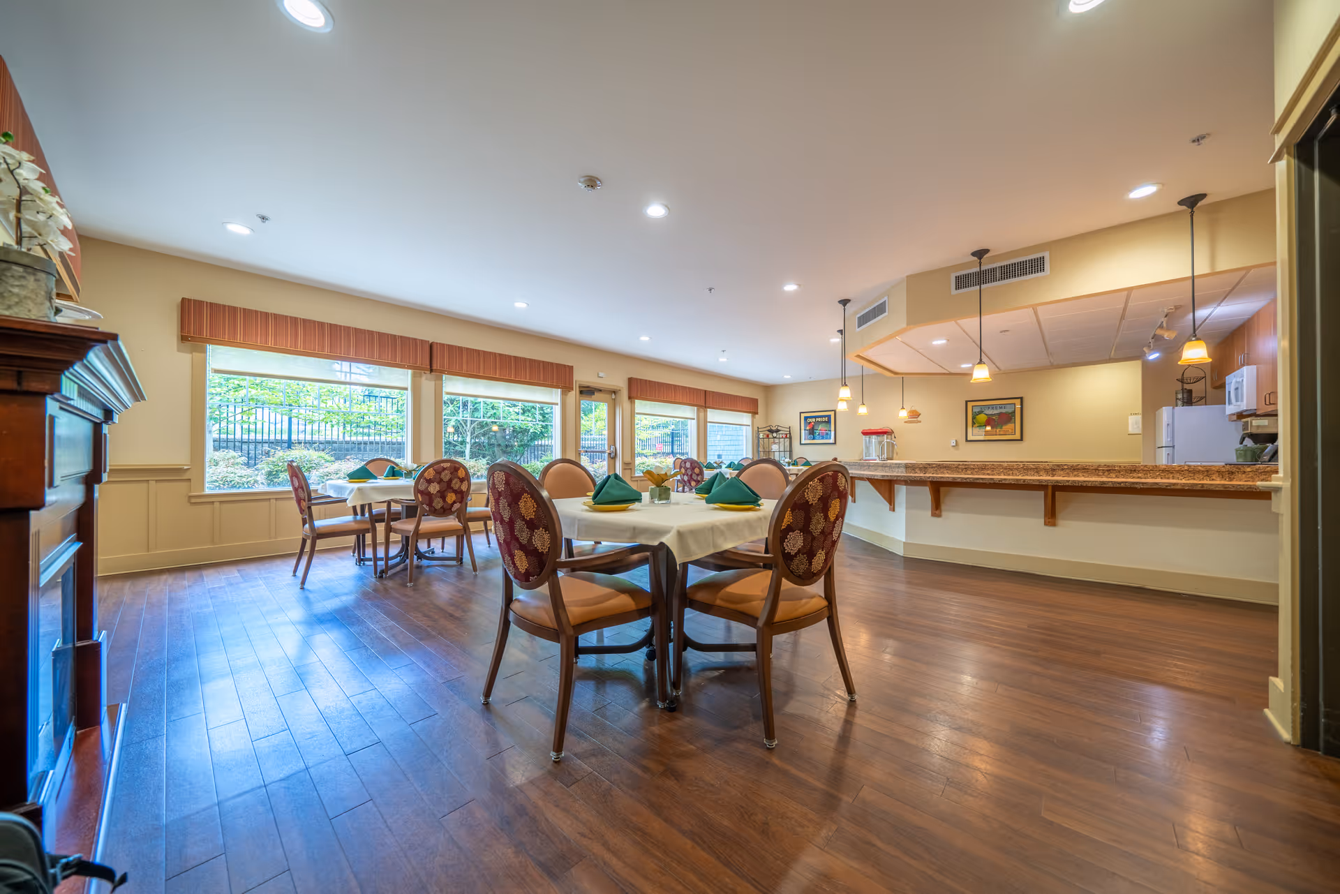 A spacious dining room with wooden flooring and large windows letting in natural light. Several round tables are set with white tablecloths, green folded napkins, and chairs with patterned upholstery. There is a long counter with pendant lights hanging above it, and a kitchen area visible in the background. The room has a warm and inviting atmosphere with light-colored walls and framed artwork.