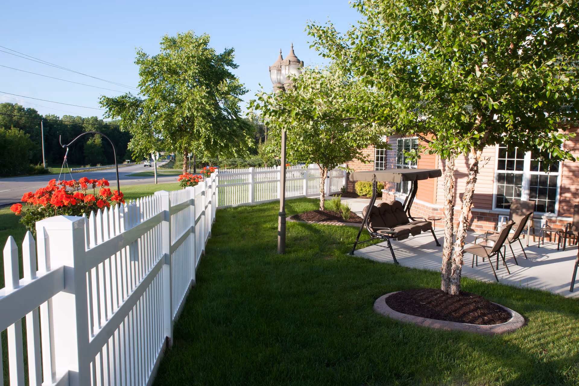 Sunny fenced garden patio with a white picket fence, hanging red flowers, trees, and outdoor seating including a swing by a brick building.