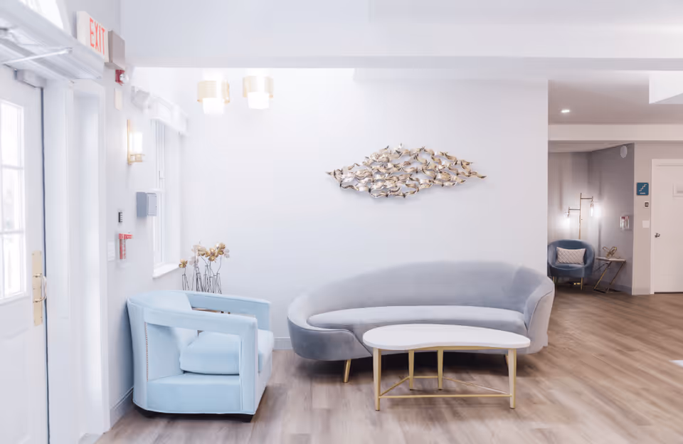 A bright and modern seating area in a senior living facility with a light blue armchair, a curved gray sofa, and a white coffee table with gold legs. The walls are white with a decorative metal wall art piece shaped like a school of fish. The floor is wood, and there are two pendant lights hanging from the ceiling. In the background, there is another blue chair and a small side table with a lamp.