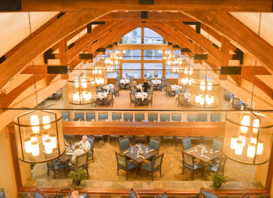 Interior view of a spacious dining room with wooden beams on the ceiling and multiple round tables set with chairs. Several pendant lights hang from the ceiling, illuminating the area. A few elderly people are seated at some of the tables, engaged in conversation.