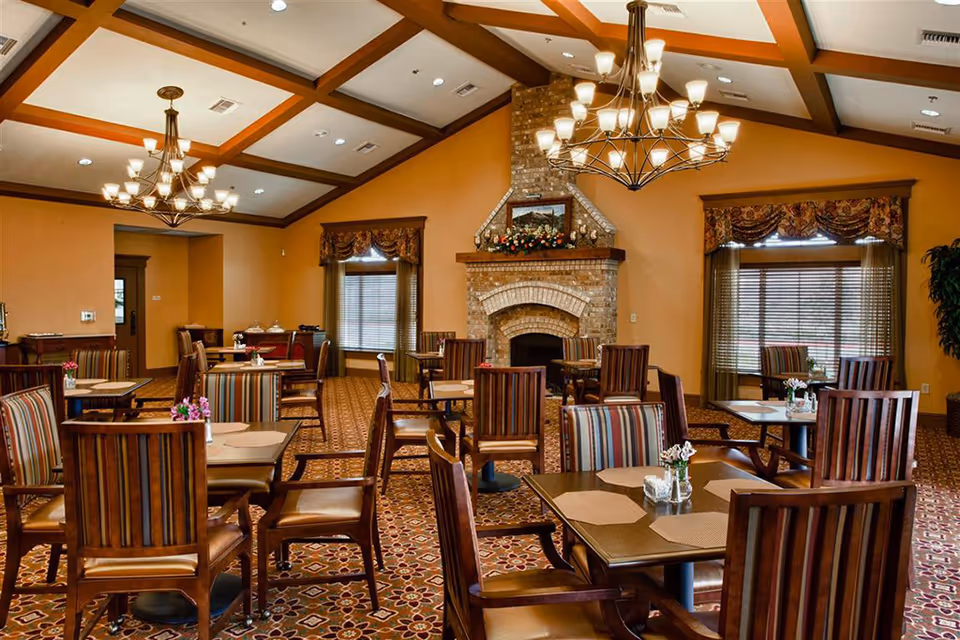 A warm dining room with multiple wooden tables and striped cushioned chairs arranged neatly. The room features a patterned carpet, two large chandeliers hanging from a coffered ceiling, and a brick fireplace decorated with flowers and a framed picture above it. Windows with blinds and valances allow natural light into the space.