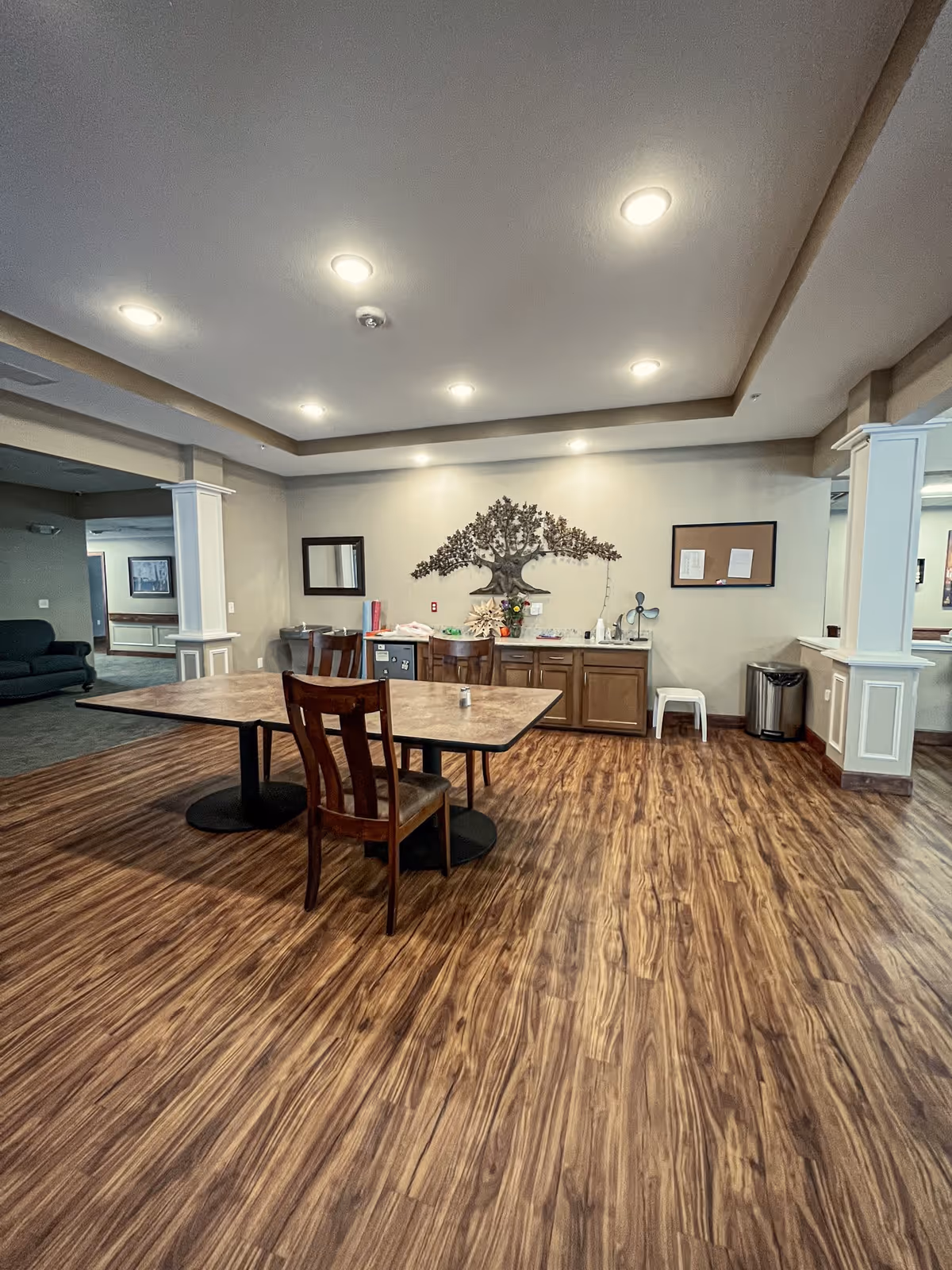 A spacious room with wood-patterned flooring and a rectangular table with four wooden chairs. The back wall features a decorative metal tree artwork above a countertop with cabinets. The ceiling has recessed lighting, and there are white pillars with molding details on either side of the room.
