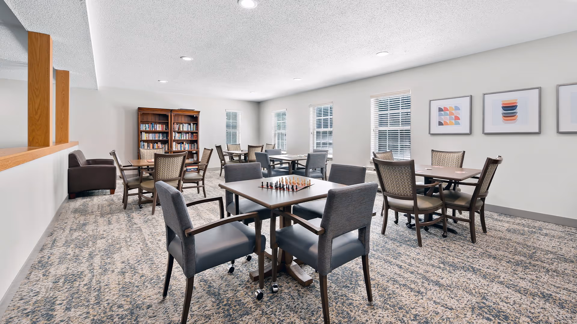 A bright and spacious common area with multiple tables and chairs arranged for group activities or dining. One table has a chessboard set up. The room has large windows with blinds, a bookshelf filled with books, and framed abstract artwork on the walls. The floor is carpeted with a patterned design, and the ceiling has recessed lighting.