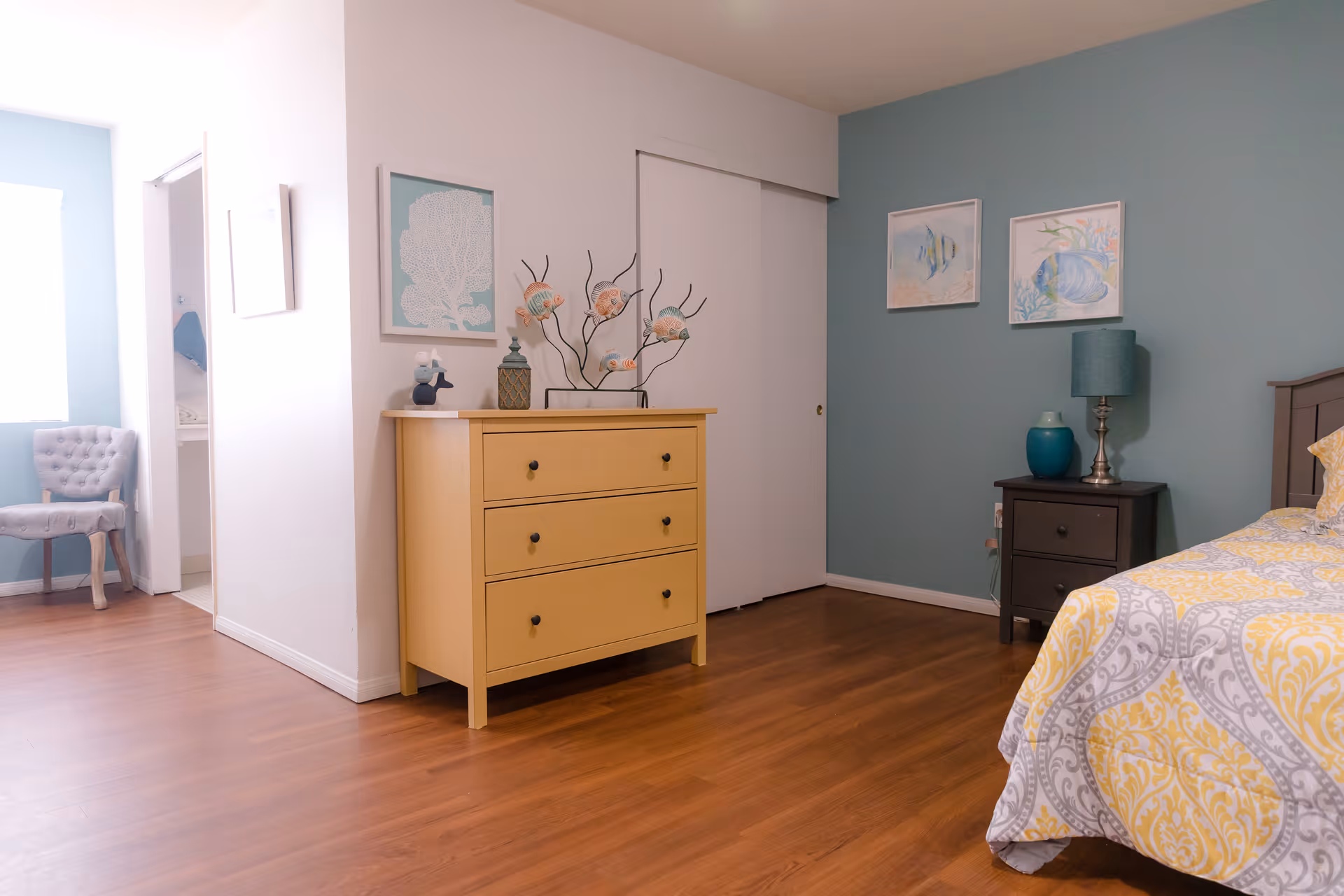 A cozy bedroom with wooden flooring, a yellow three-drawer dresser adorned with decorative items, a nightstand with a lamp and vase, a bed with a yellow and gray patterned bedspread, and blue walls decorated with framed artwork. A gray upholstered chair is visible near a window in an adjacent room.
