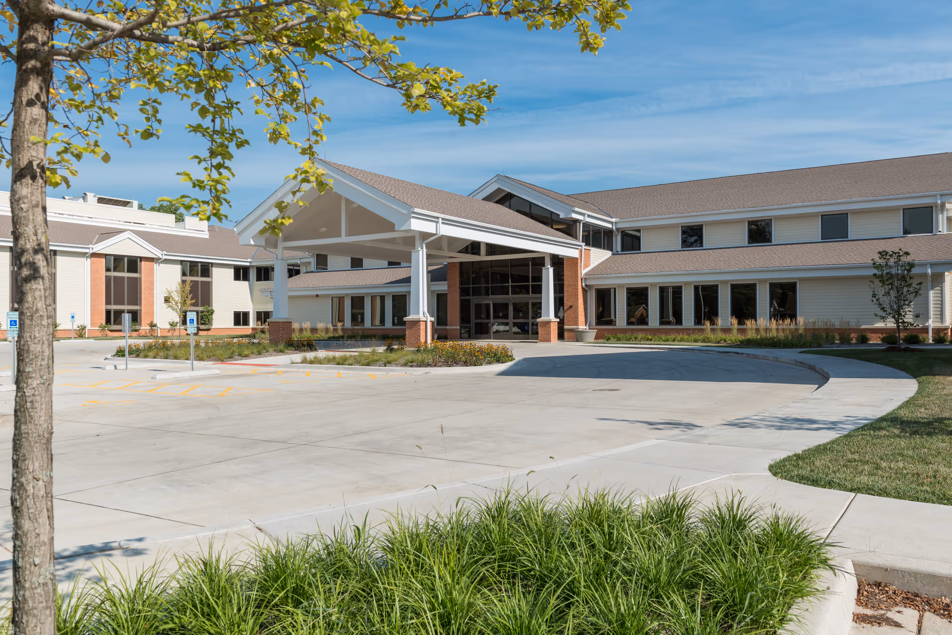 Exterior view of a senior living facility with a covered entrance, large windows, and a circular driveway surrounded by landscaped greenery and trees under a clear blue sky.