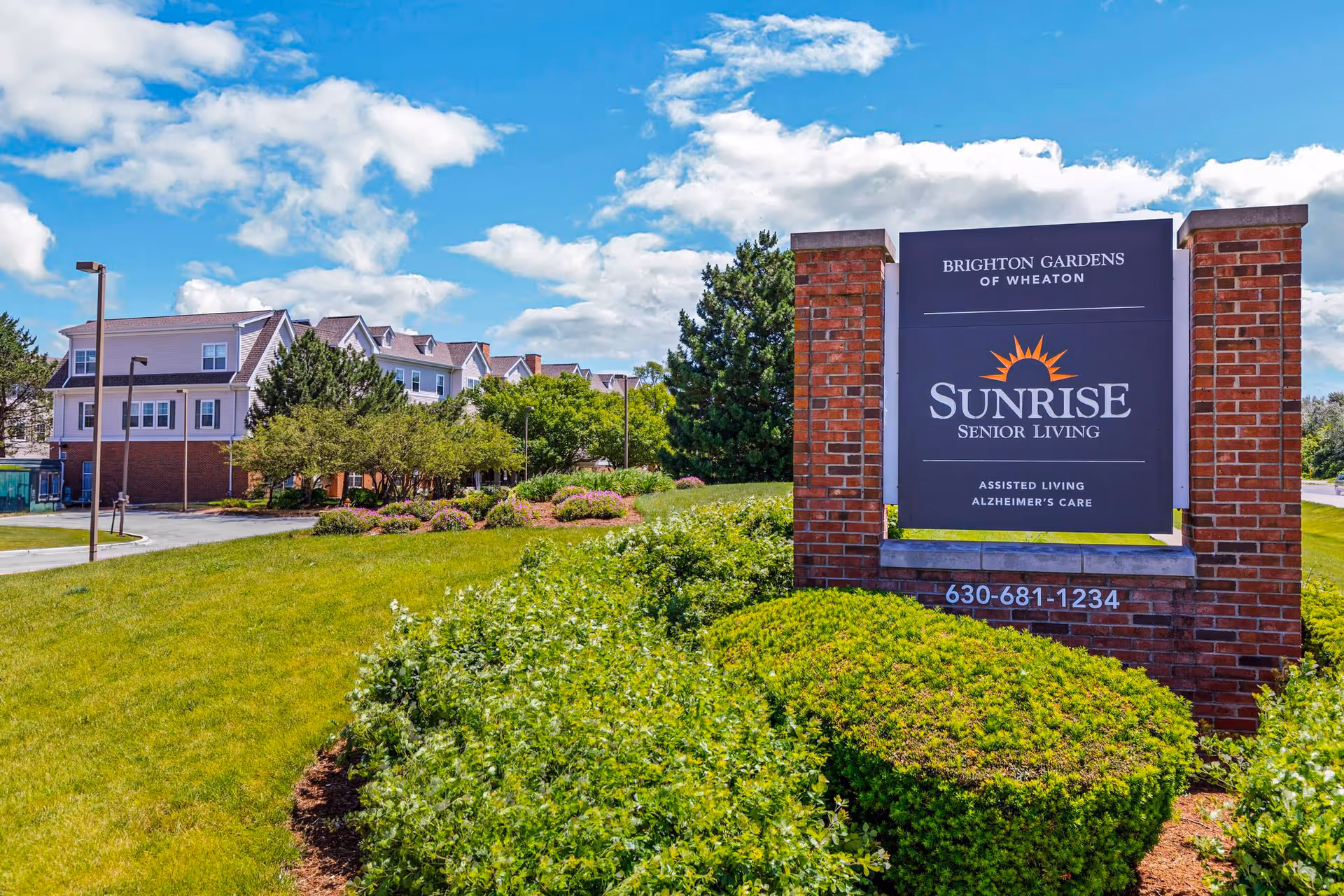 Exterior view of Brighton Gardens of Wheaton senior living facility with a large sign displaying Sunrise Senior Living, assisted living, and Alzheimer's care. The building is surrounded by green grass, bushes, trees, and a partly cloudy blue sky.