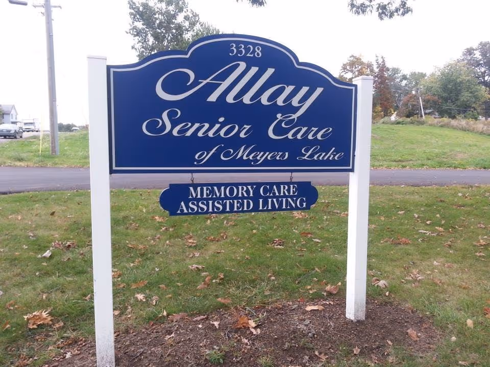 A blue and white outdoor sign for Allay Senior Care of Meyers Lake, indicating services for memory care and assisted living, situated on a grassy area near a road with trees and houses in the background.