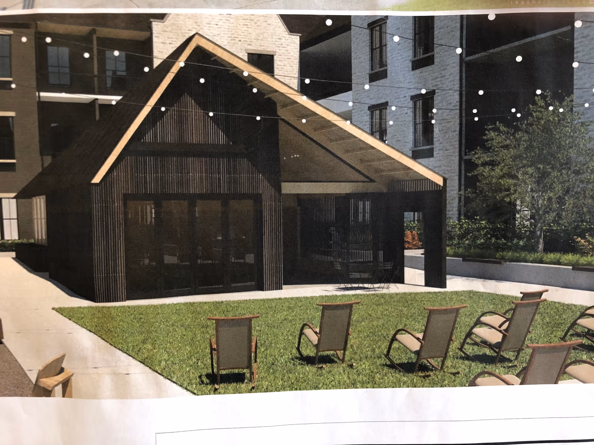 Outdoor seating area with multiple chairs arranged on a grassy lawn in front of a modern pavilion structure with a peaked roof. String lights are hung above the area, and there are buildings and trees in the background.