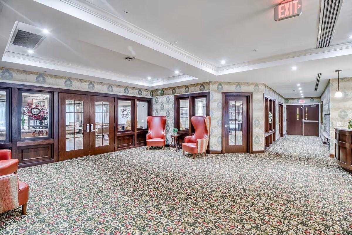 A spacious, well-lit interior common area with patterned carpet and wallpaper. The room features dark wood-framed glass doors, two red upholstered armchairs with a small table between them, and ceiling lights. Exit signs are visible above two doorways.
