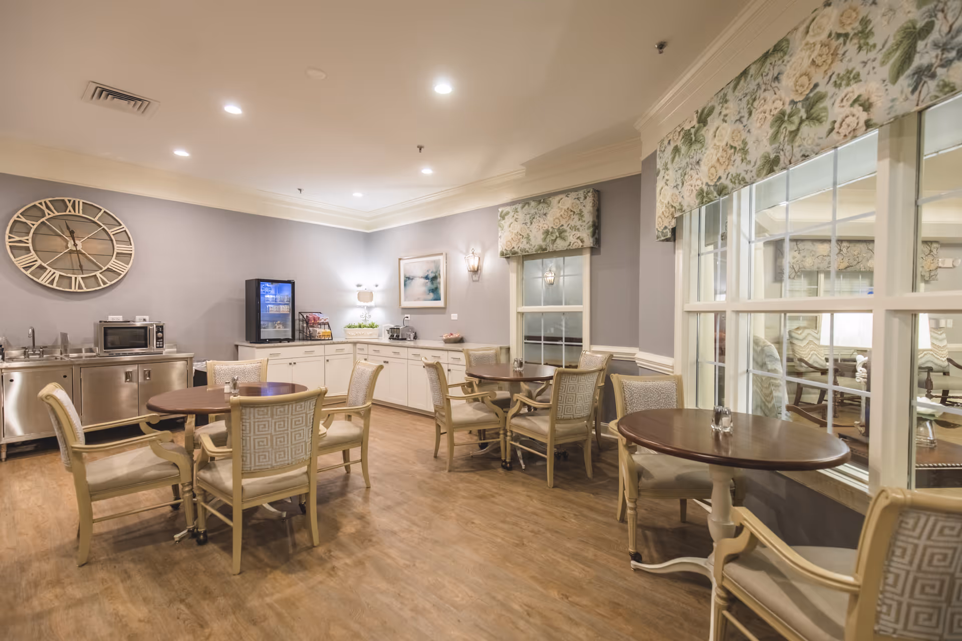 A cozy dining area in a senior living facility with round wooden tables and cushioned chairs. The room features light gray walls, floral valances over the windows, a large decorative wall clock, and a kitchenette area with a microwave, mini fridge, and coffee station. The floor is wood, and the space is softly lit with ceiling lights and wall sconces.