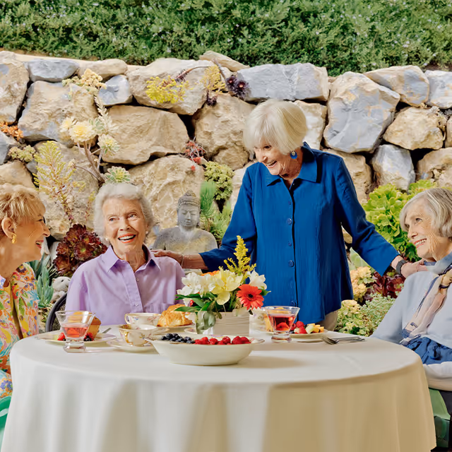 Four elderly women enjoying a cheerful conversation around a round table set with tea cups, plates of fruit, and pastries in an outdoor garden area with a stone wall and greenery in the background.