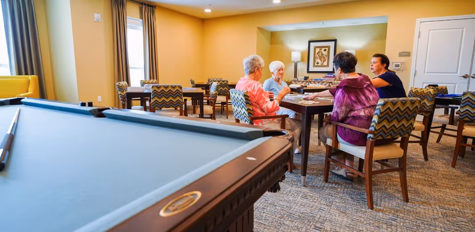 A group of four elderly women sitting around a table playing a game in a well-lit common room with a pool table in the foreground and several tables and chairs in the background.