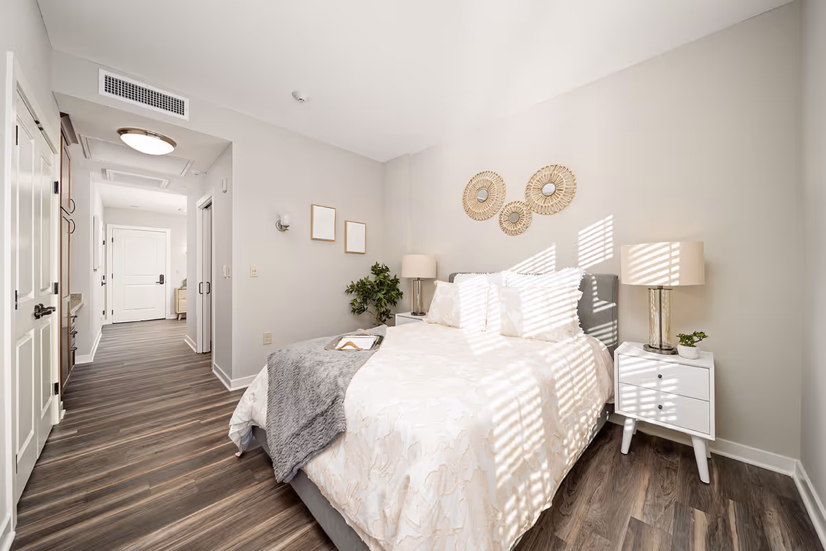 A bright and modern bedroom with a large bed covered in white and beige bedding and a gray throw blanket. There are two white nightstands on either side of the bed, each with a lamp and a small plant. The walls are light-colored with three decorative round mirrors above the bed and two small framed pictures on the adjacent wall. The floor is wood with a dark finish, and there is a hallway leading to a white door in the background.