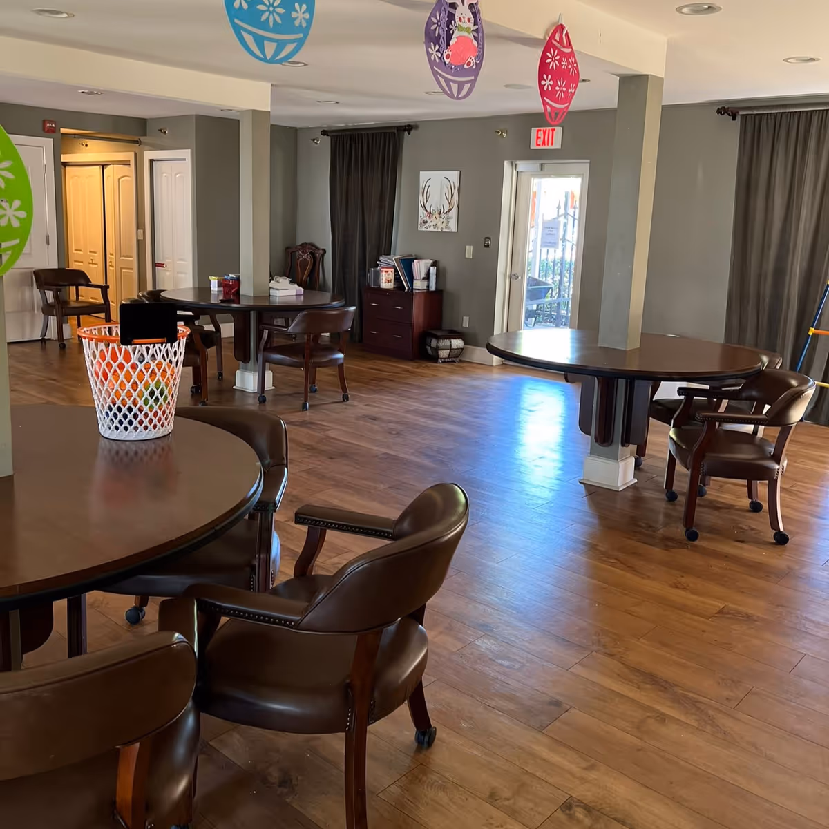 Interior view of a common area in an assisted living facility with round wooden tables and brown leather chairs on wheels. The room has wood flooring, gray walls, and dark curtains covering windows. Colorful hanging decorations are visible from the ceiling, and there is a door with an exit sign above it. A small cabinet with books and other items is against one wall.