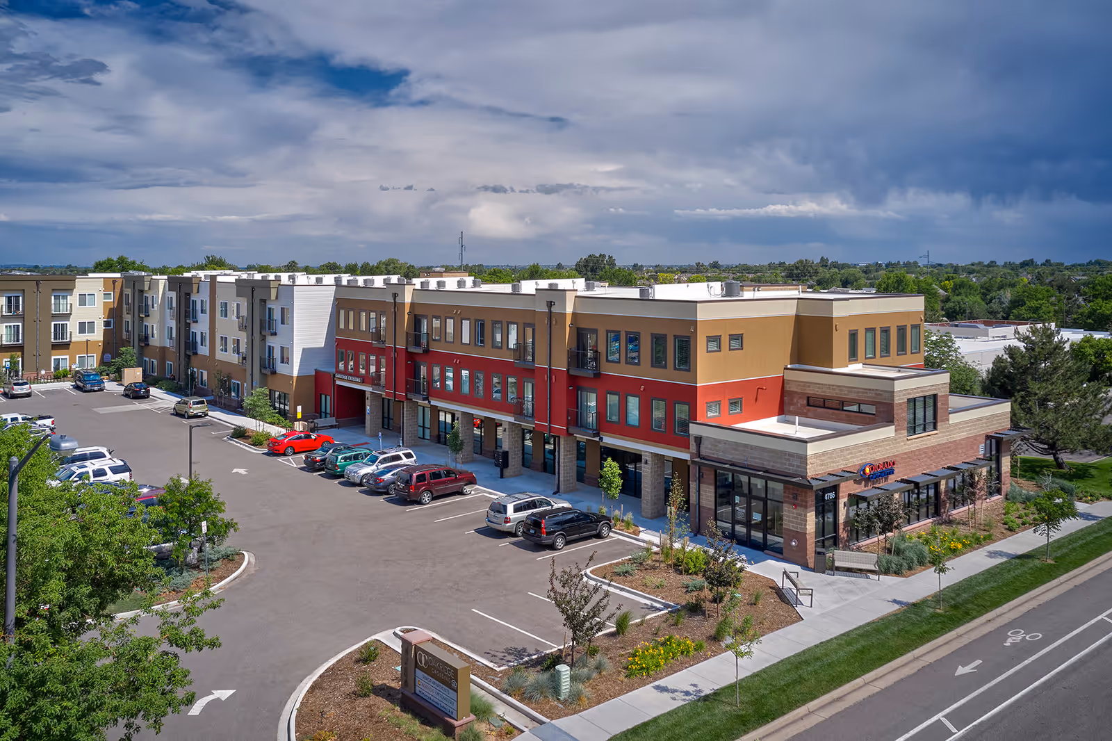 Exterior view of Oakridge Crossing, a modern three-story senior living facility with a parking lot in front. The building features a mix of beige, brown, and red colors with multiple windows and balconies. There are several cars parked in the lot, landscaped areas with trees and shrubs, and a clear sky with some clouds above.