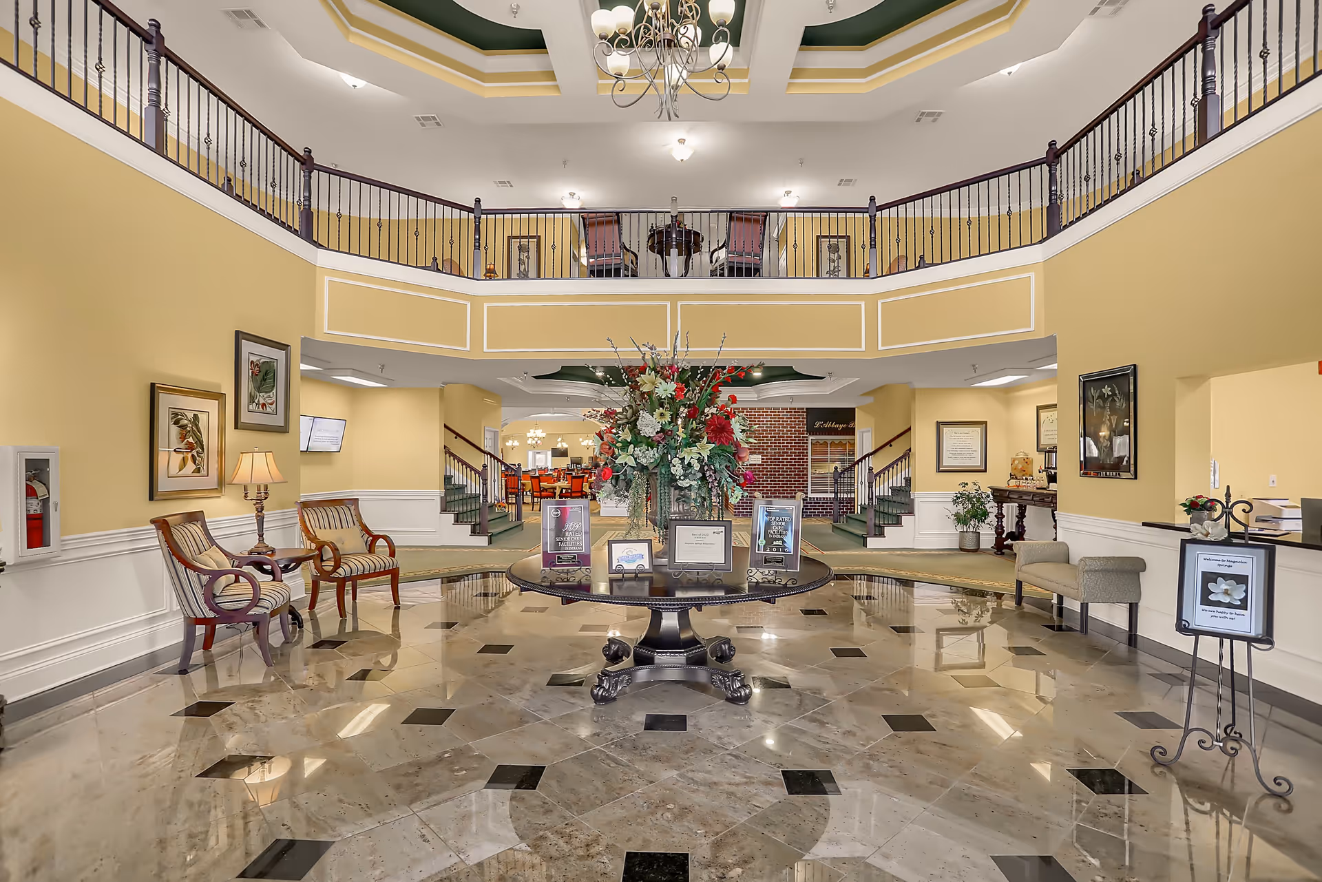 Spacious and elegant senior living facility lobby with polished marble floor, a round table with a large floral arrangement in the center, seating areas with chairs and benches along the walls, a chandelier hanging from a decorative ceiling, and a balcony overlooking the lobby.