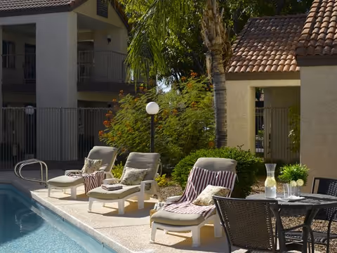 Poolside courtyard with lounge chairs, a small table holding a pitcher and glasses, and apartment-style buildings and palm trees in the background.